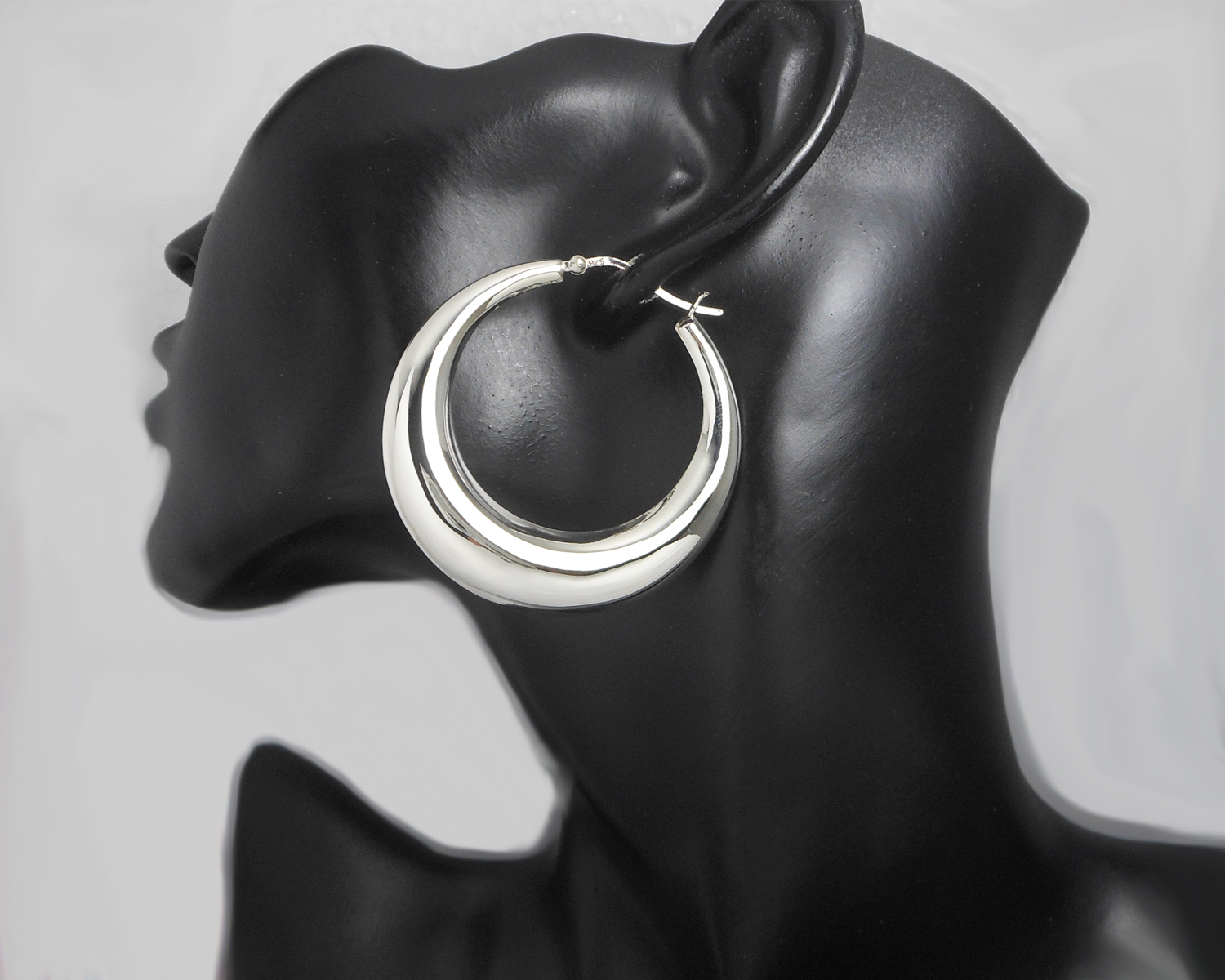 A pair of large, chunky 925 sterling silver puffy hoop earrings (50mm) shown on a black mannequin display and laid out on a clean surface.