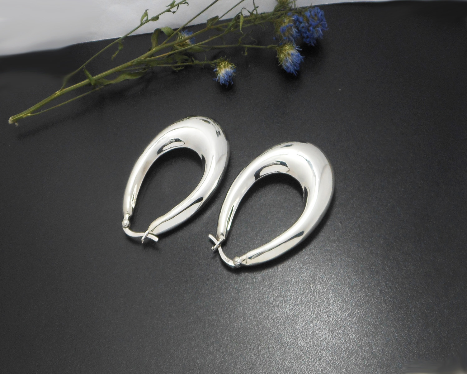 A pair of handcrafted 925 sterling silver puffy oval hoop earrings (47mm) resting on a rustic wood log slice next to a small bunch of dried blue flowers.