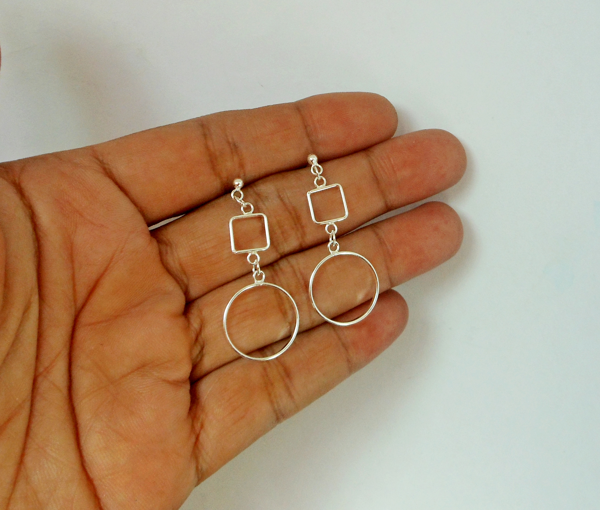 A pair of handcrafted 925 sterling silver drop earrings featuring an open square connected to an open circle, displayed in a hand.