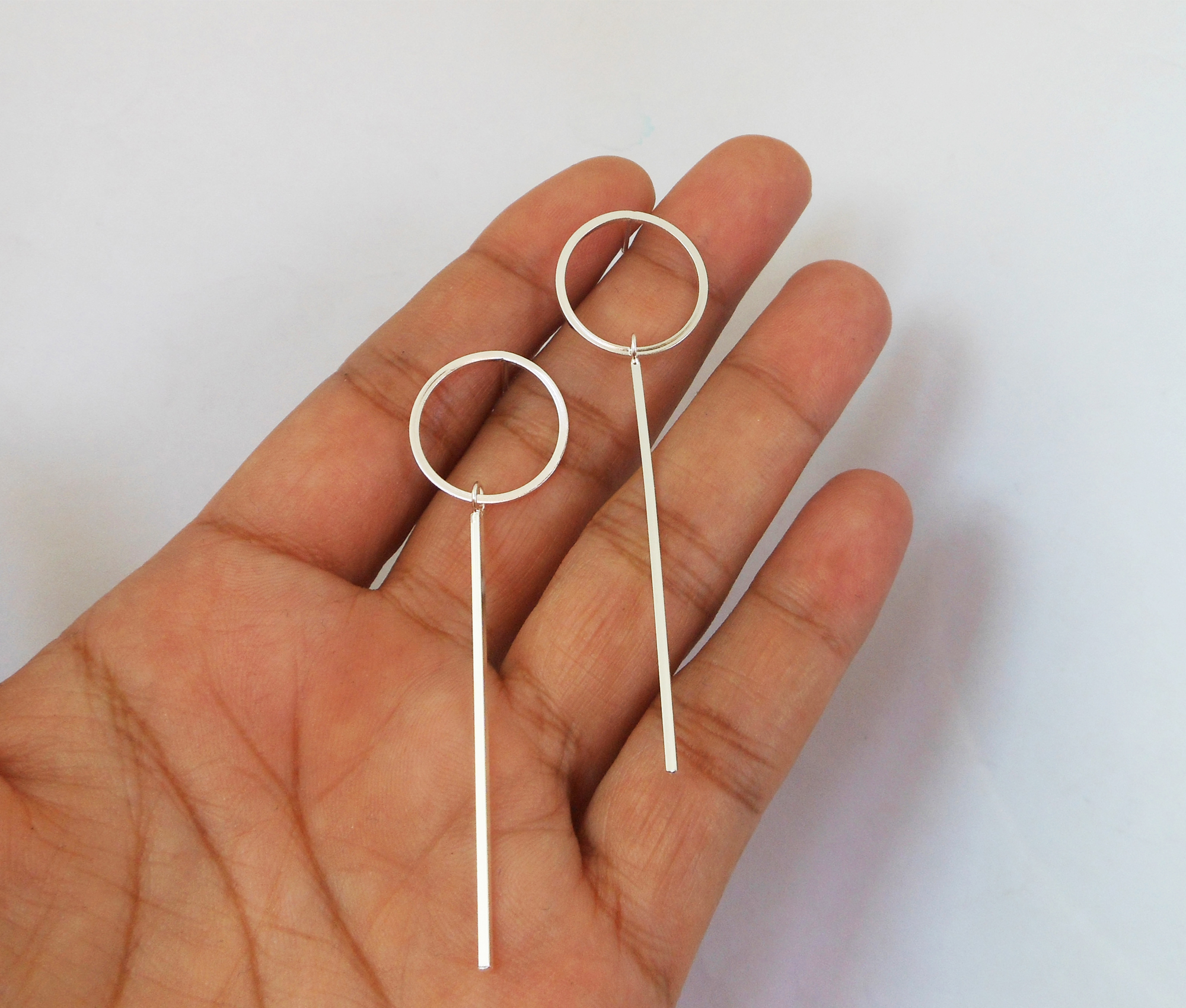 A pair of handcrafted 925 sterling silver geometric drop stud earrings featuring a clean circle loop paired with a sleek vertical bar, resting in a hand.
