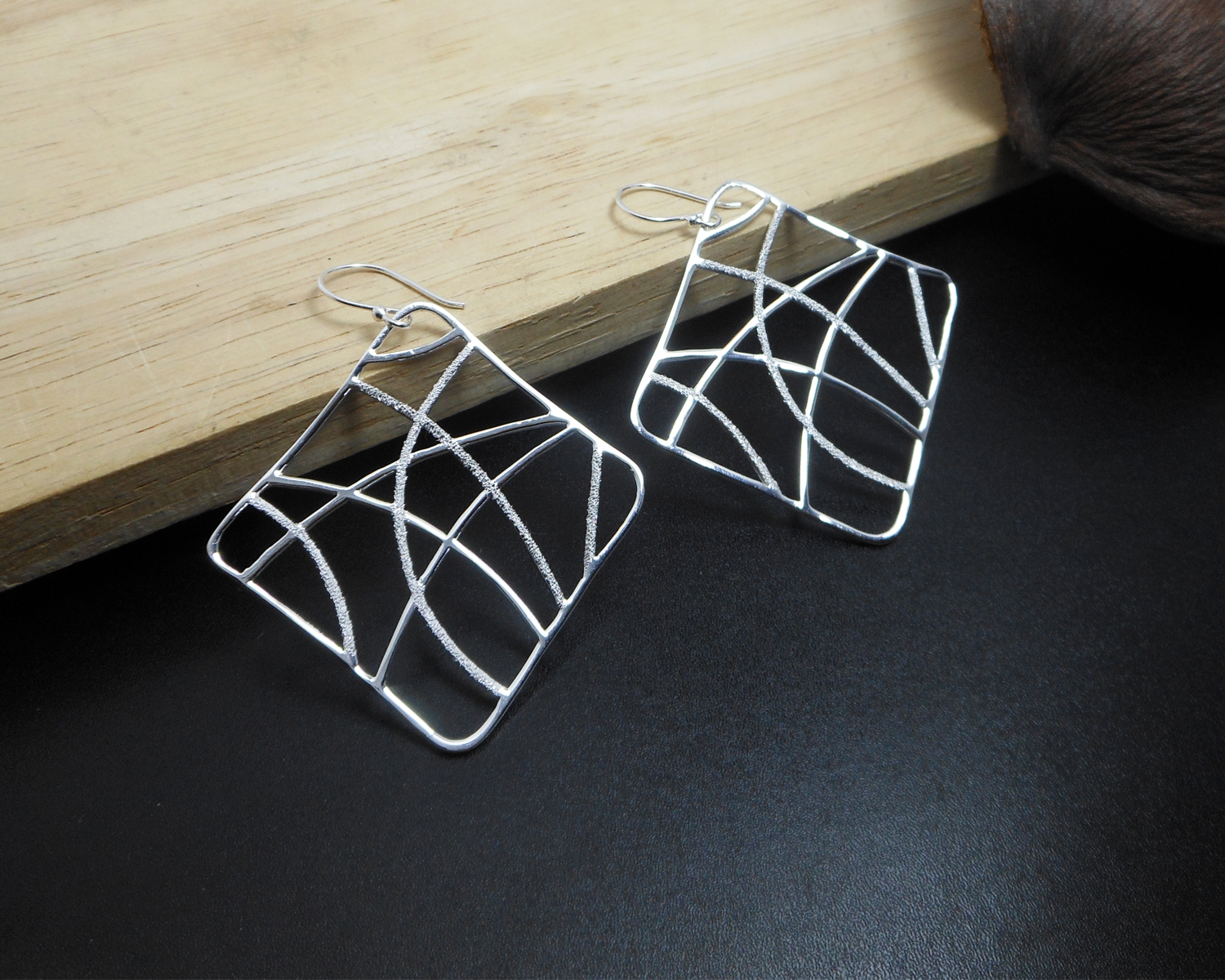 A pair of handcrafted 925 sterling silver perforated web square drop earrings featuring an abstract textured pattern on a dark surface next to a wooden block.