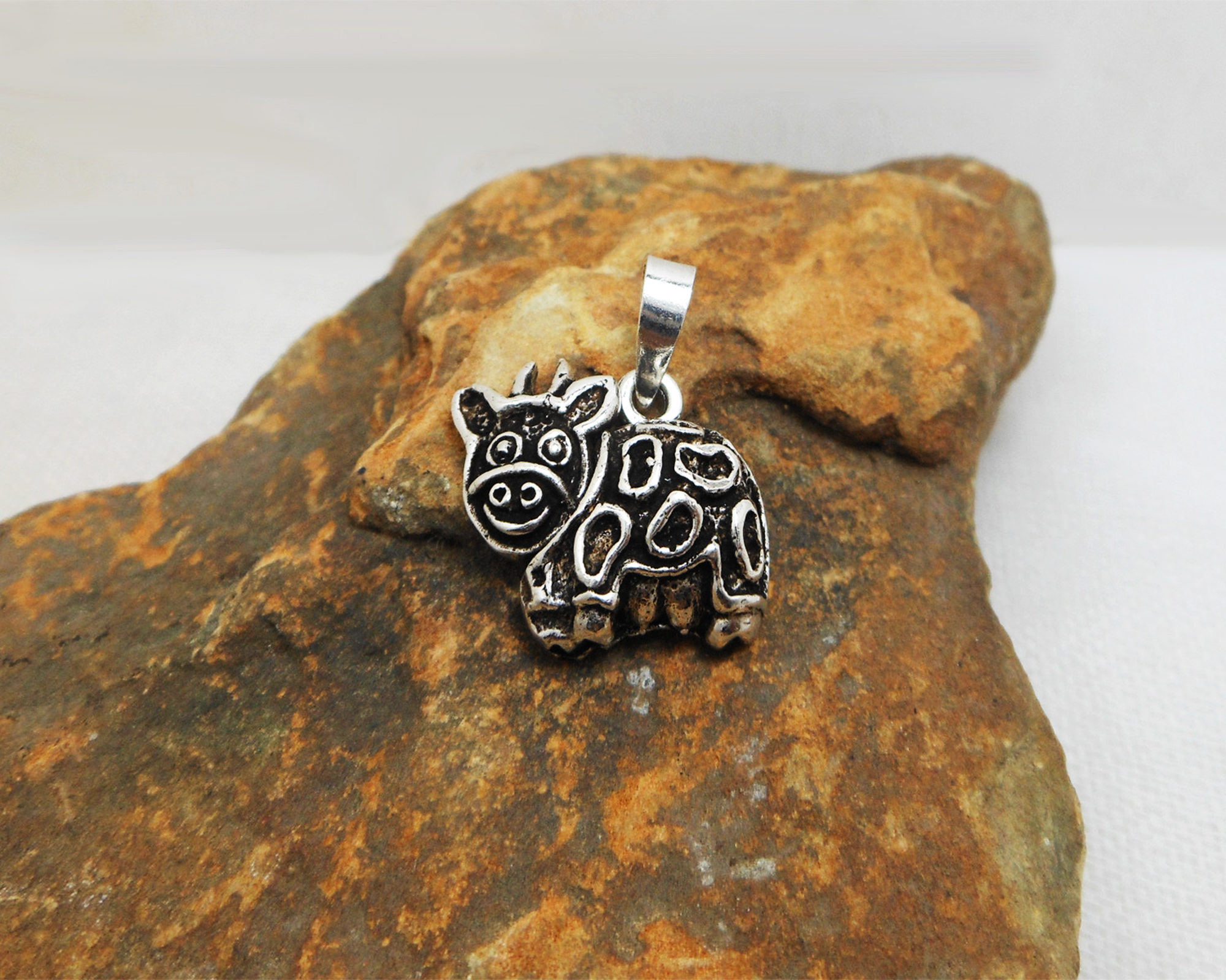 A detailed photograph of a handcrafted 925 sterling silver cow pendant with a movable design and textured finish, resting on a rustic stone surface.