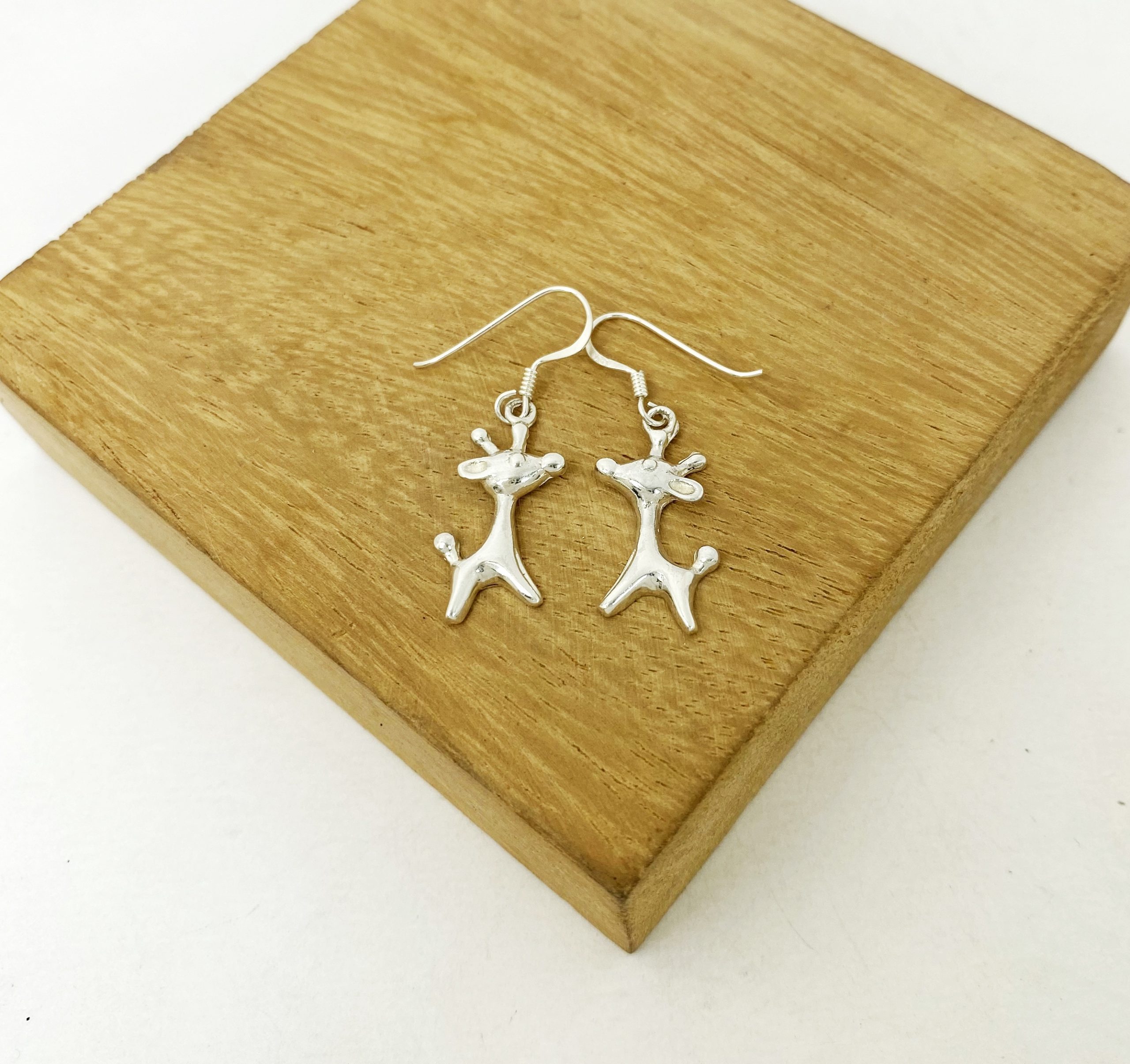 A pair of handcrafted 925 sterling silver solid giraffe drop earrings featuring a smooth, high-polished finish resting on a textured piece of wood.