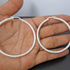 A pair of handcrafted 925 sterling silver 50mm hoop earrings held in a hand, showcasing a brilliant, intricate diamond-cut textured finish.