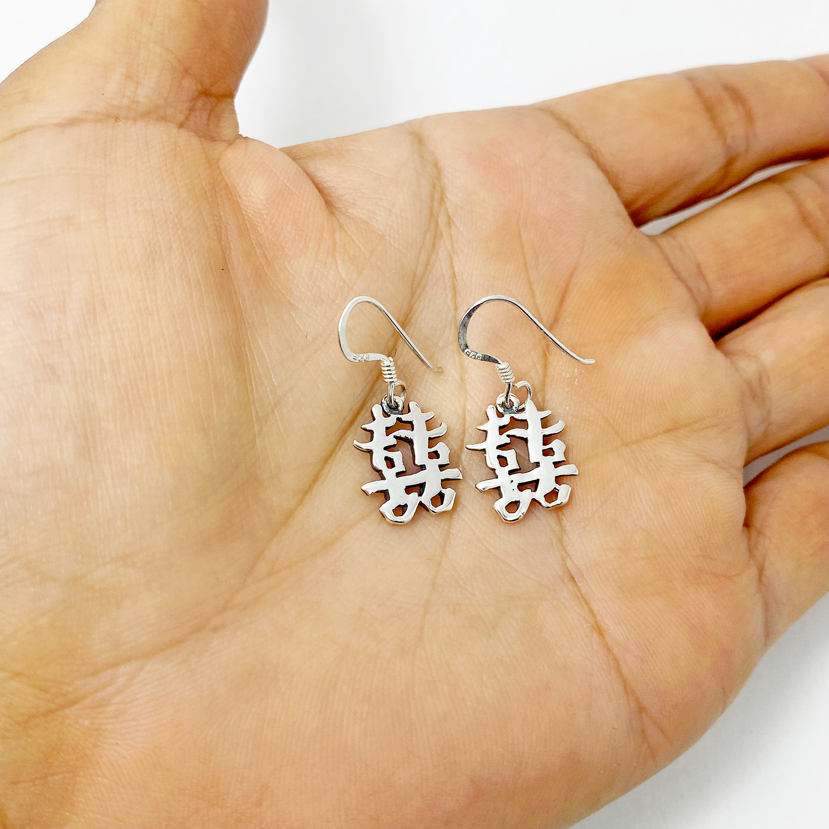 A pair of handcrafted 925 sterling silver drop earrings featuring the Chinese character for "Happiness" in a polished, solid cutout design, held in a hand.
