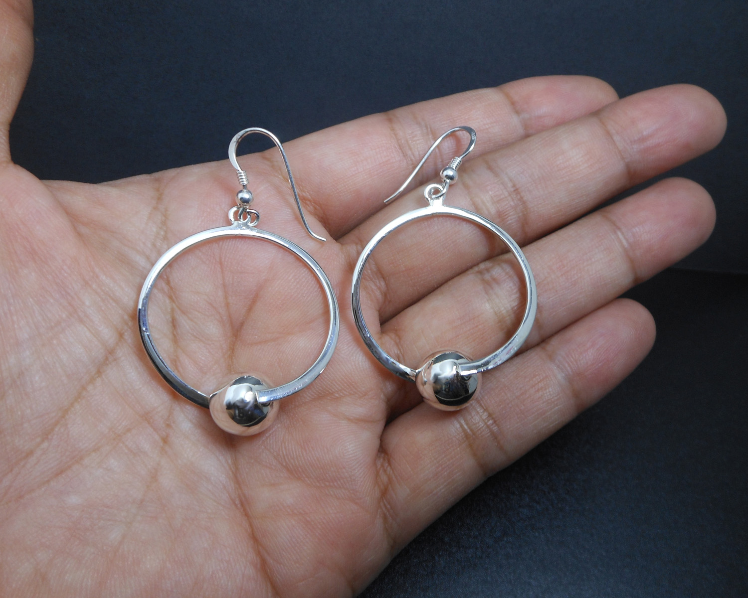 A pair of handcrafted 925 sterling silver drop earrings featuring a circular frame with a polished silver bead, displayed in a hand.