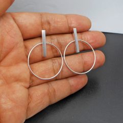 A pair of handcrafted 925 sterling silver dangle earrings featuring a minimalist circle hoop suspended from a vertical bar stud, held in a hand.