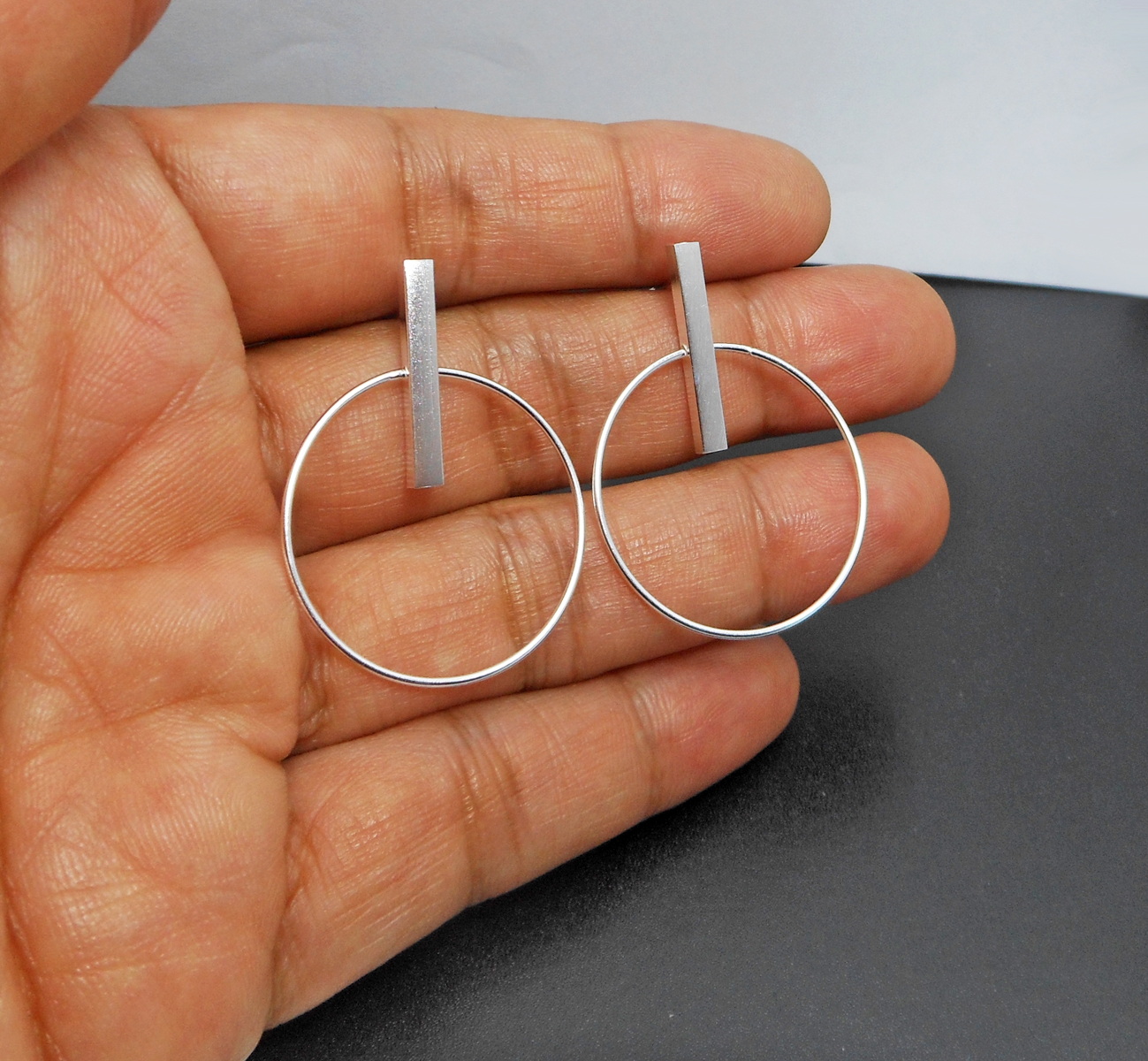 A pair of handcrafted 925 sterling silver dangle earrings featuring a minimalist circle hoop suspended from a vertical bar stud, held in a hand.