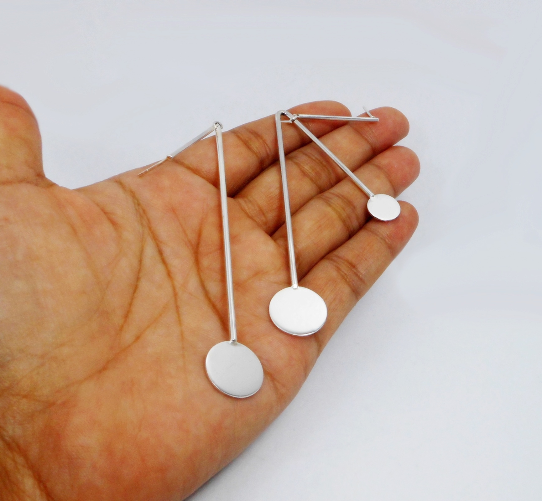 A pair of handcrafted 925 sterling silver mismatched dangle earrings featuring a modern geometric design with solid polished discs and spinning bars, held in a hand.