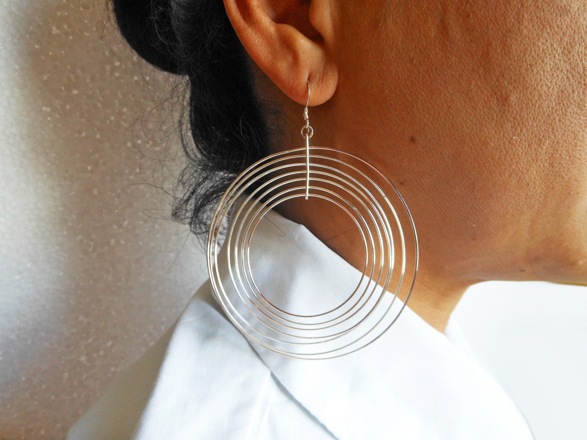 A close-up profile view of a woman wearing large, lightweight 925 sterling silver dangle earrings composed of multiple concentric, smooth, shiny, and spinning circles, referencing life style model