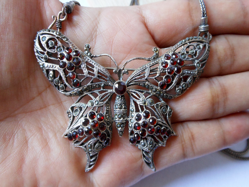 A detailed close-up of a large, handcrafted 925 sterling silver butterfly pendant adorned with intricate marcasite stones and deep red garnet accents, designed to be worn as both a brooch and a necklace set.