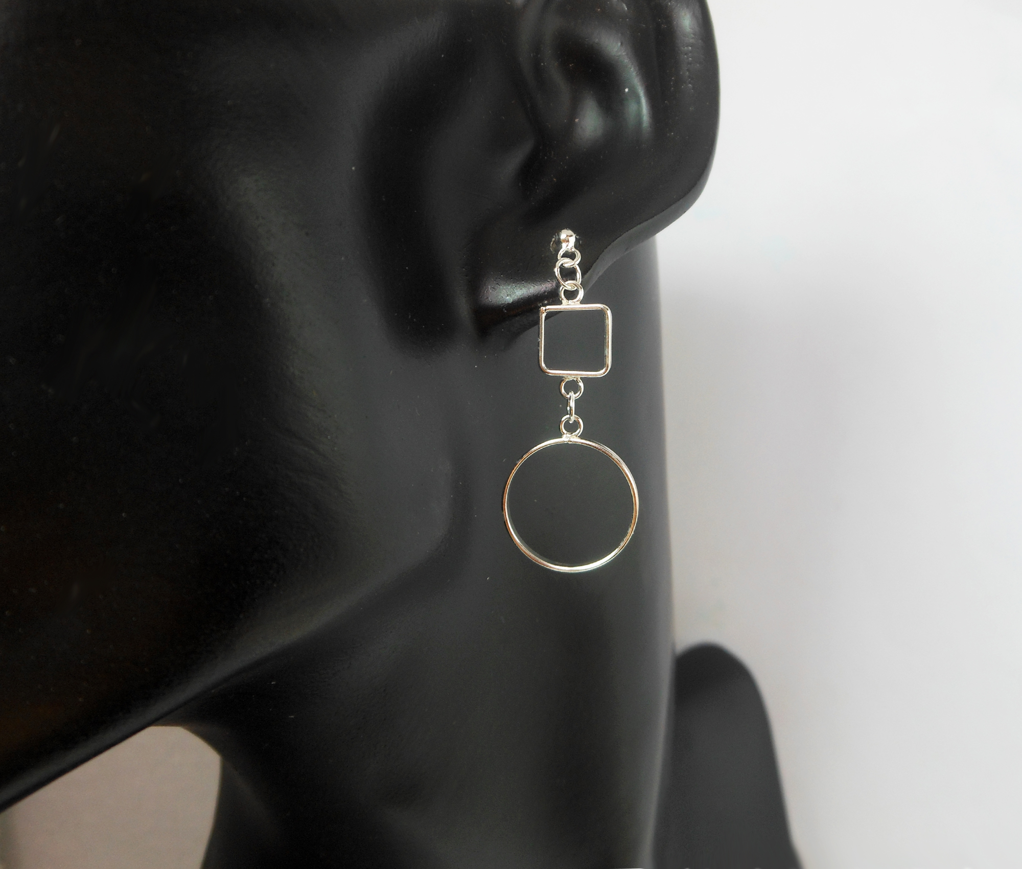 A pair of handcrafted 925 sterling silver drop earrings featuring an open square connected to an open circle, displayed on a black mannequin ear.