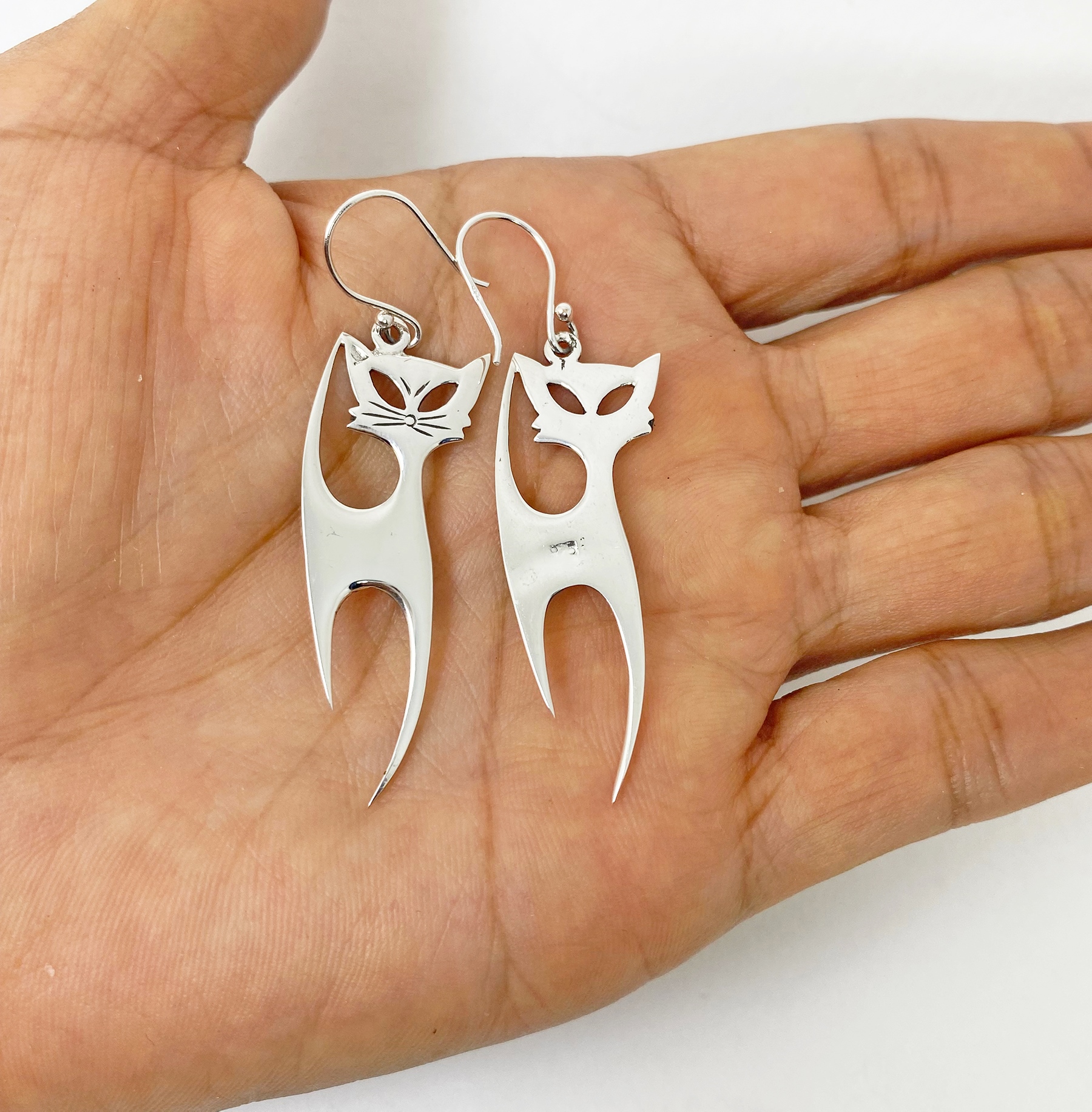 A pair of handcrafted 925 sterling silver long cat drop earrings featuring an engraved feline silhouette with a high-polished finish, held in a hand.