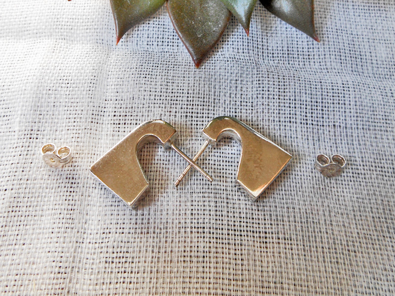 Pair of handmade 925 sterling silver puffy geometric stud earrings with a sleek, architectural design, resting on a textured fabric surface.