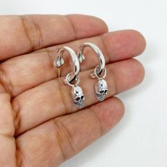 A pair of handcrafted 925 sterling silver hoop earrings featuring a unique bone-shaped hoop and a dangling skull charm, held in a hand.