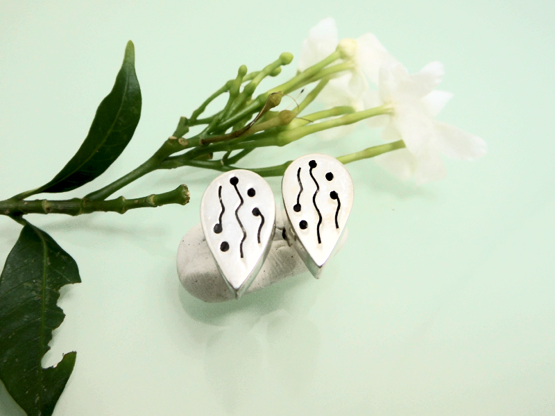 Pair of handmade 925 sterling silver teardrop stud earrings featuring a unique etched wavy line and dot pattern with a puffy, dimensional profile, resting near white flowers.