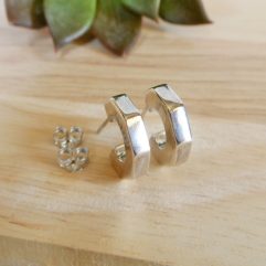 A pair of small, hand-polished 925 sterling silver hoop earrings with a unique puffy, multi-faceted pentagonal shape, resting on a light wood surface with a blurred succulent in the background.