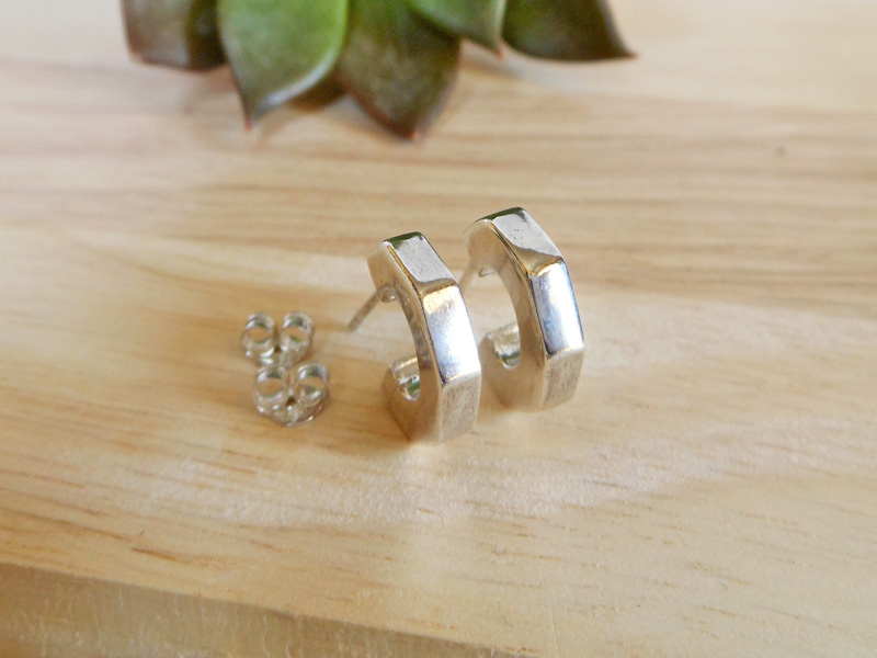 A pair of small, hand-polished 925 sterling silver hoop earrings with a unique puffy, multi-faceted pentagonal shape, resting on a light wood surface with a blurred succulent in the background.