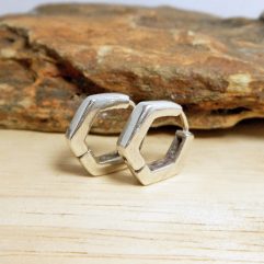 A pair of handcrafted 925 sterling silver pentagonal huggie hoop earrings resting on a piece of natural wood.