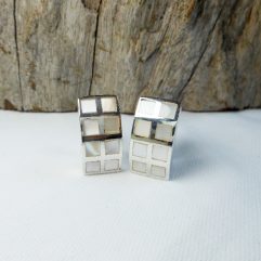A pair of handcrafted 925 sterling silver rectangular hoop earrings, featuring inlaid square mother of pearl tiles with a grid pattern, resting on a white surface in front of a piece of weathered wood.