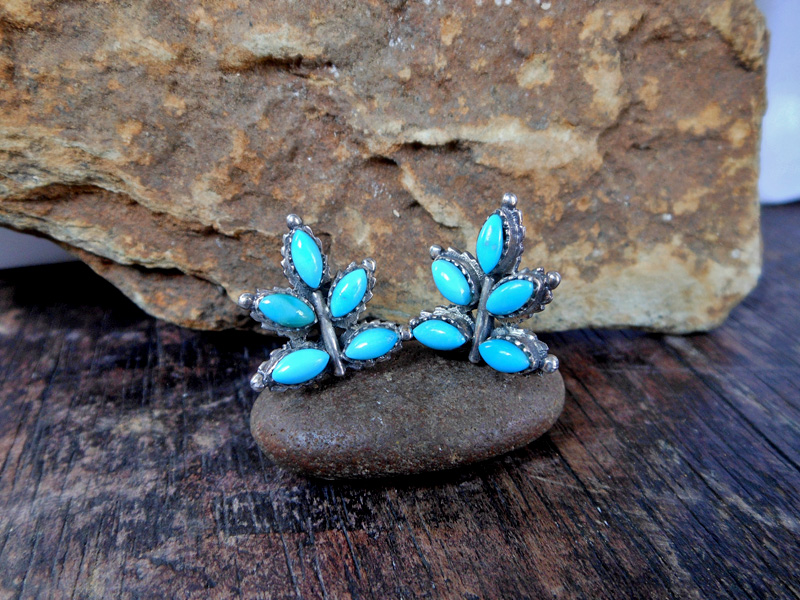 A pair of handcrafted 925 sterling silver stud earrings, shaped like a cluster of leaves, set with small, oval-cut natural blue turquoise stones, resting on a small smooth stone.