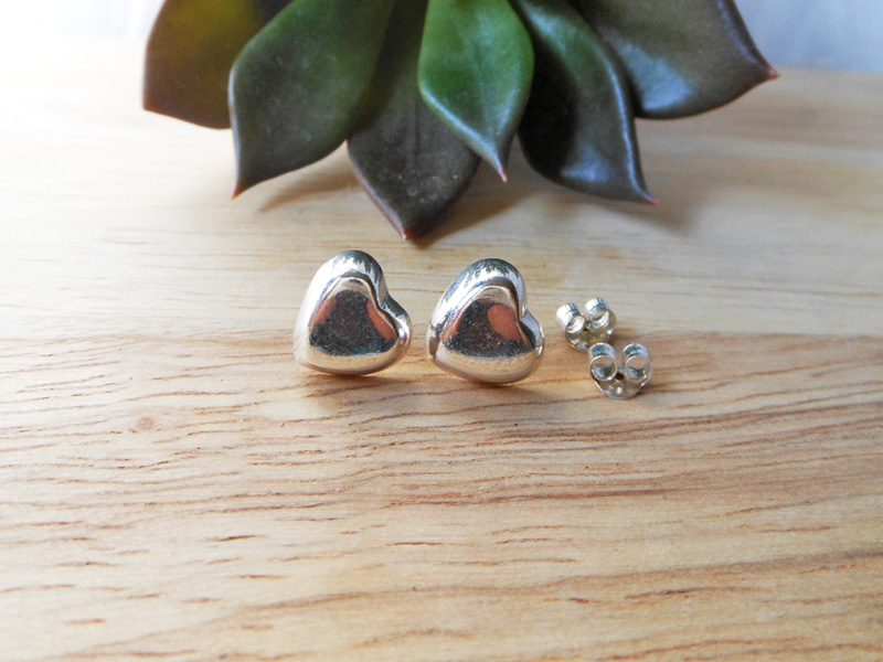 Pair of handmade 925 sterling silver puffy heart stud earrings resting on a wooden surface with a succulent in the background.