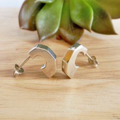 Pair of handmade 925 sterling silver puffy half hoop stud earrings with a sleek geometric design, resting on a wooden surface with a succulent in the background.