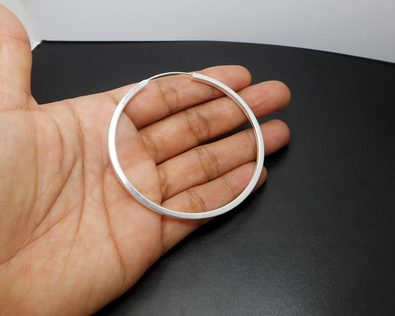 A pair of large, 65mm handmade 925 sterling silver hoop earrings featuring a unique square tube profile held in a hand