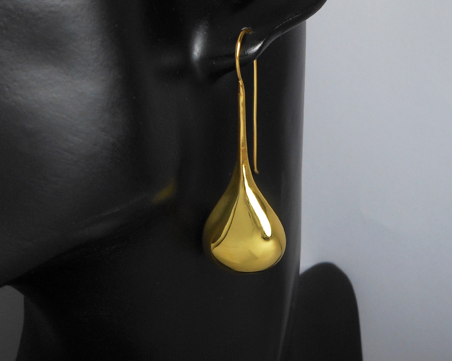 A long, gold-plated teardrop threader earring shown on a dark display stand.