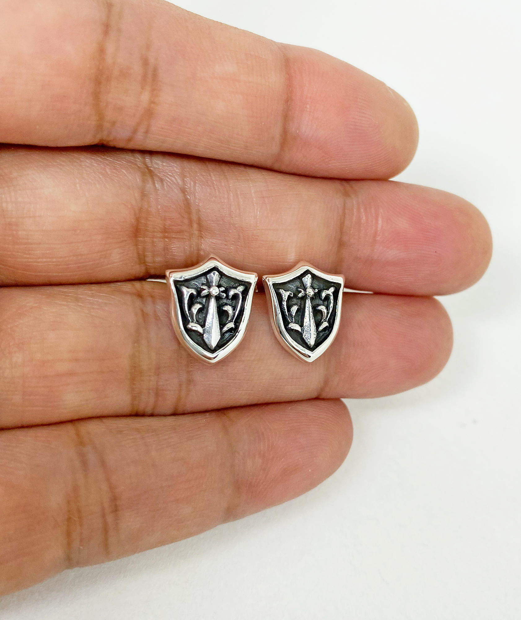 Close-up of sterling silver sword and shield stud earrings held in a hand.