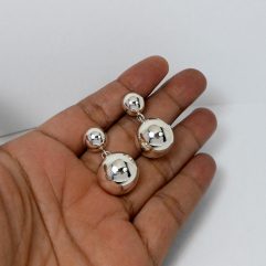 A pair of handcrafted 925 sterling silver earrings featuring a smaller ball stud and a larger ball charm drop, displayed in a hand