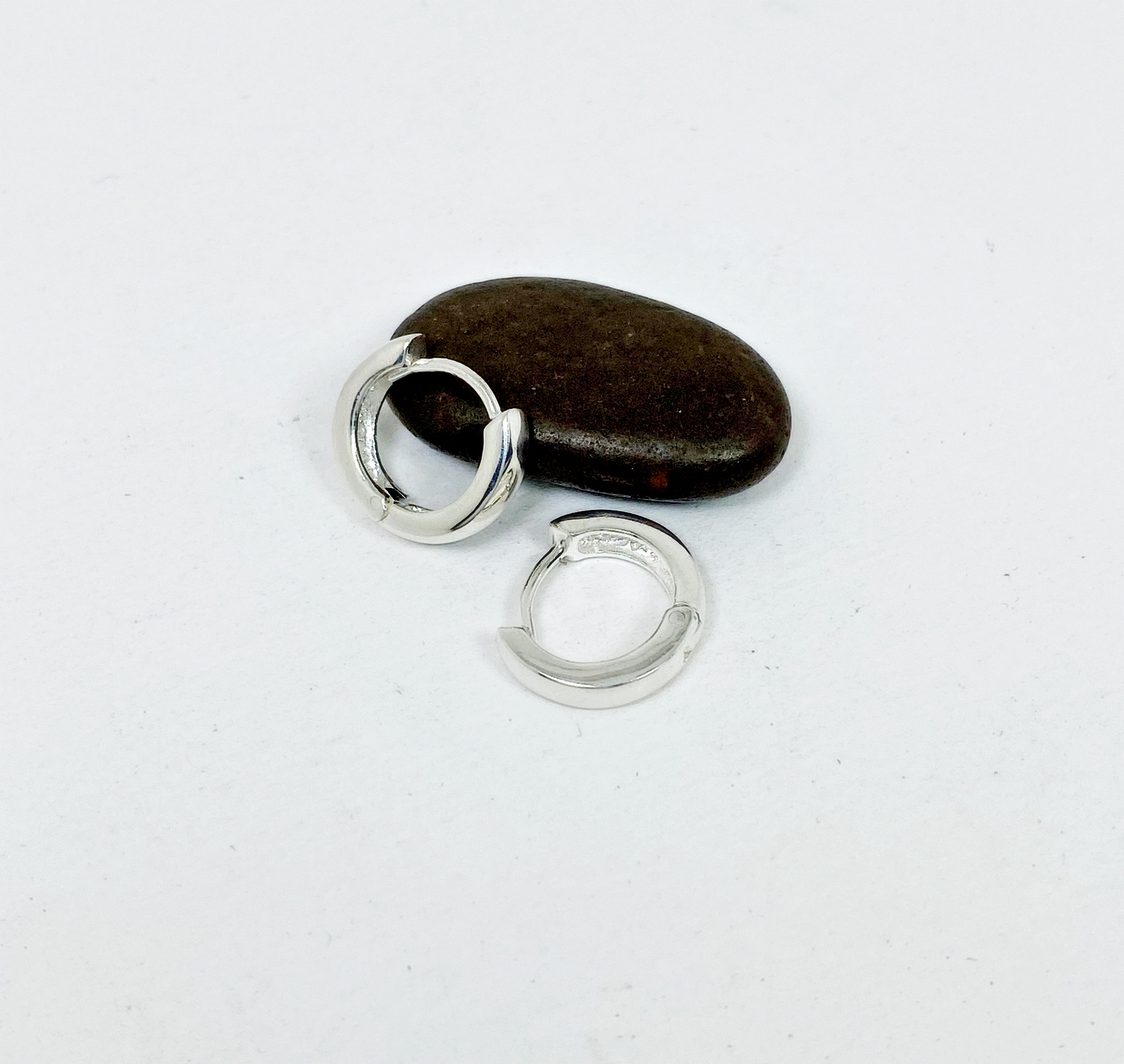 A pair of polished 925 sterling silver round huggie hoop earrings, 3mm wide, resting next to a smooth dark stone.