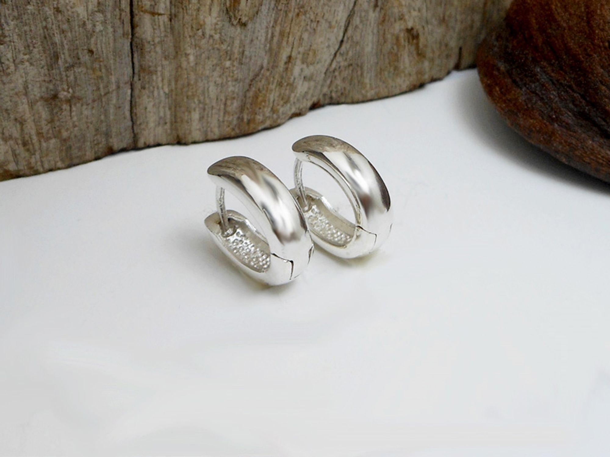 A pair of polished 925 sterling silver oval huggie hoop earrings, 4mm wide, resting on a white surface with wooden textures in the background.