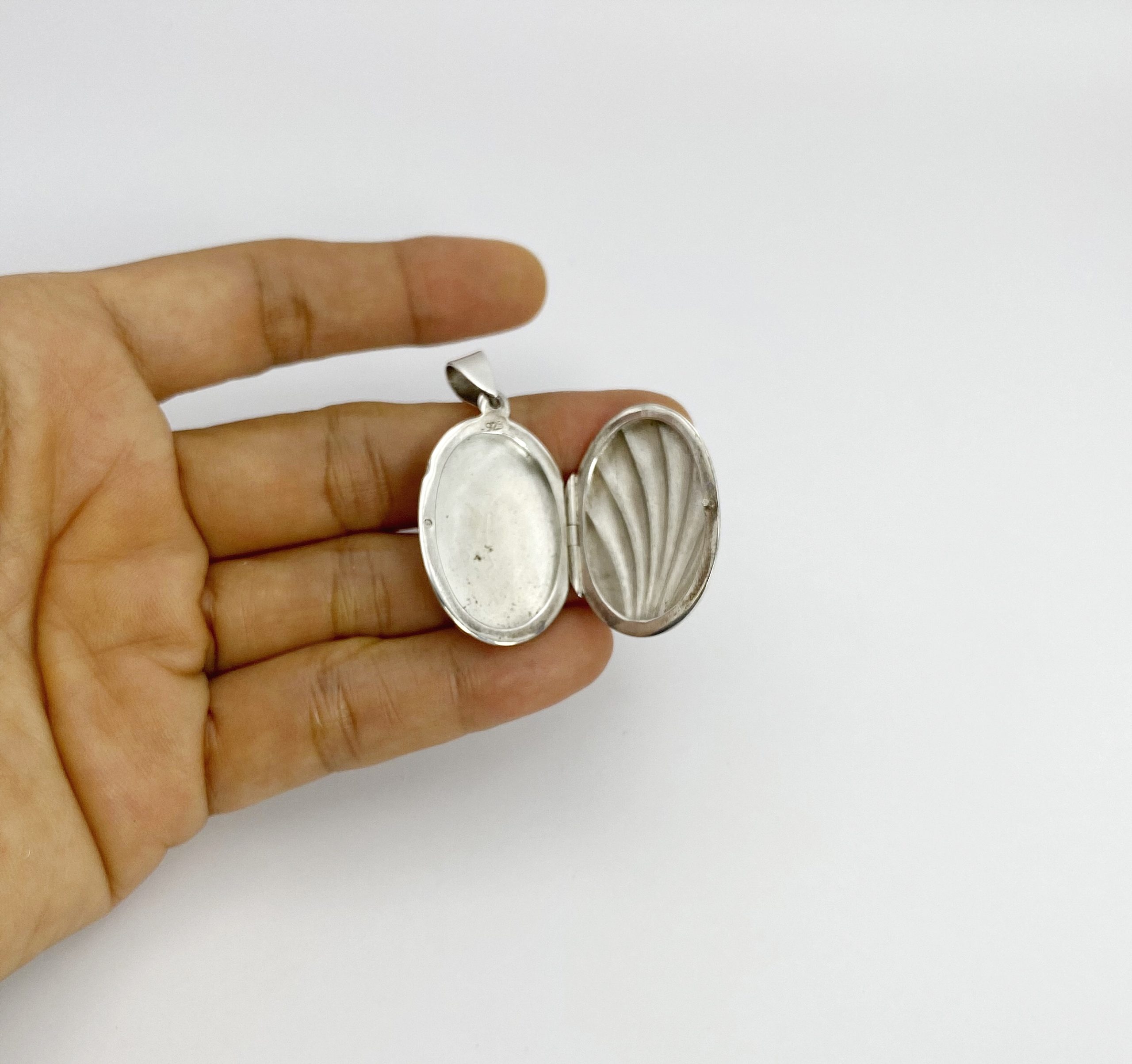 Hand holding an open oxidized sterling silver shell locket displaying the internal photo slot and decorative ribbing texture inside.