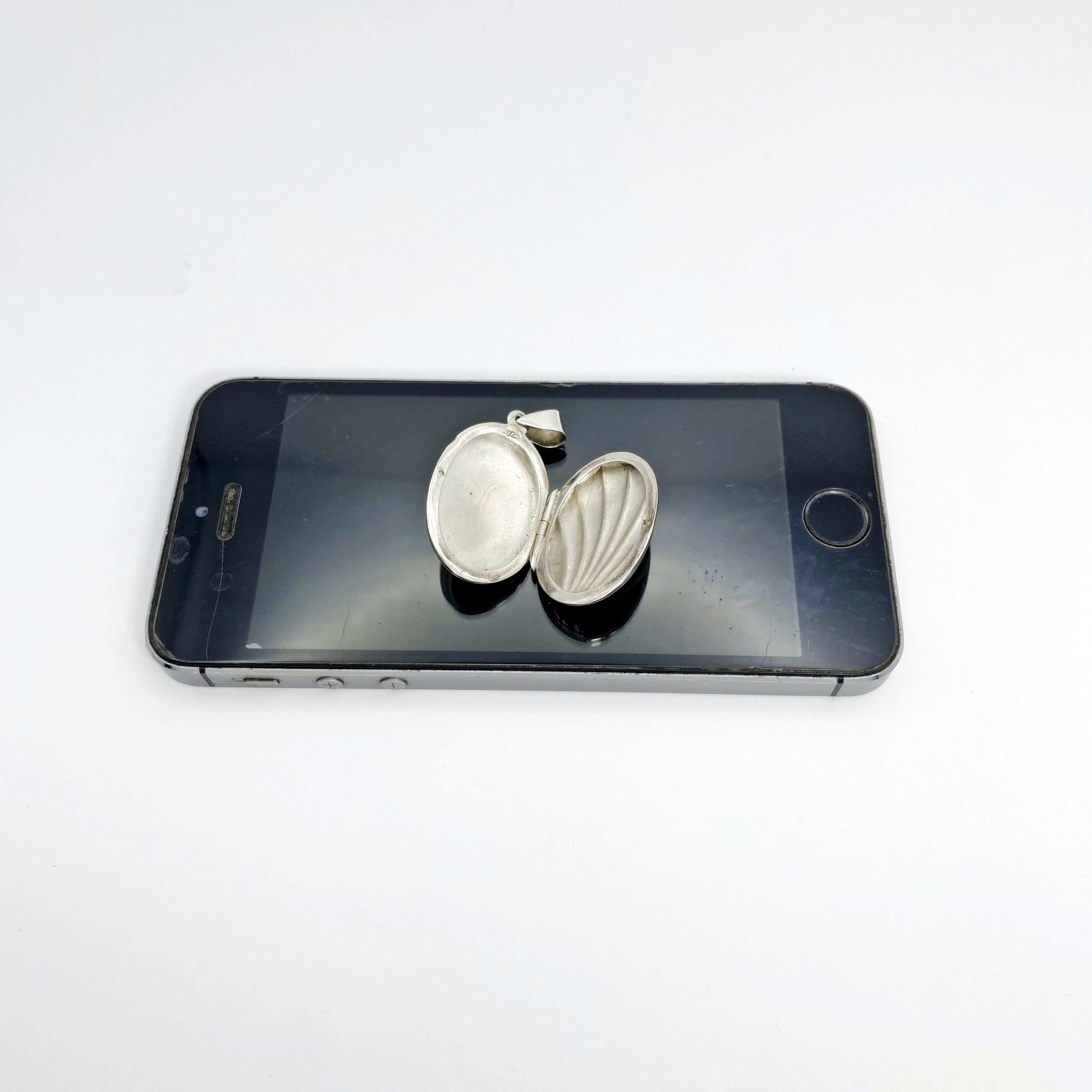 Top view of an open oxidized silver shell locket resting on a mobile phone screen, showcasing the internal photo slots and detailed ribbing texture.