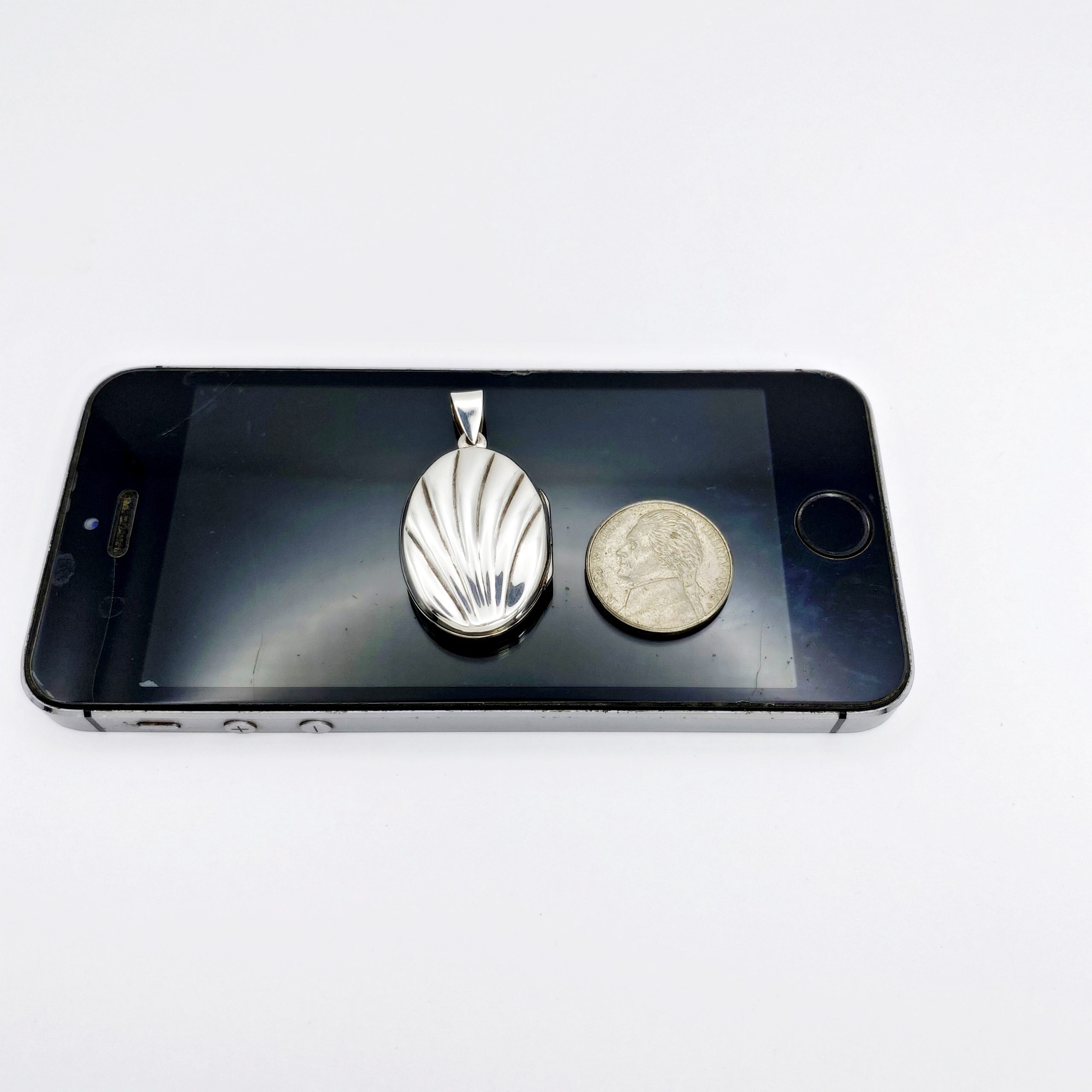 Size comparison view of an oxidized sterling silver shell locket pendant placed next to a coin to demonstrate its actual product scale.
