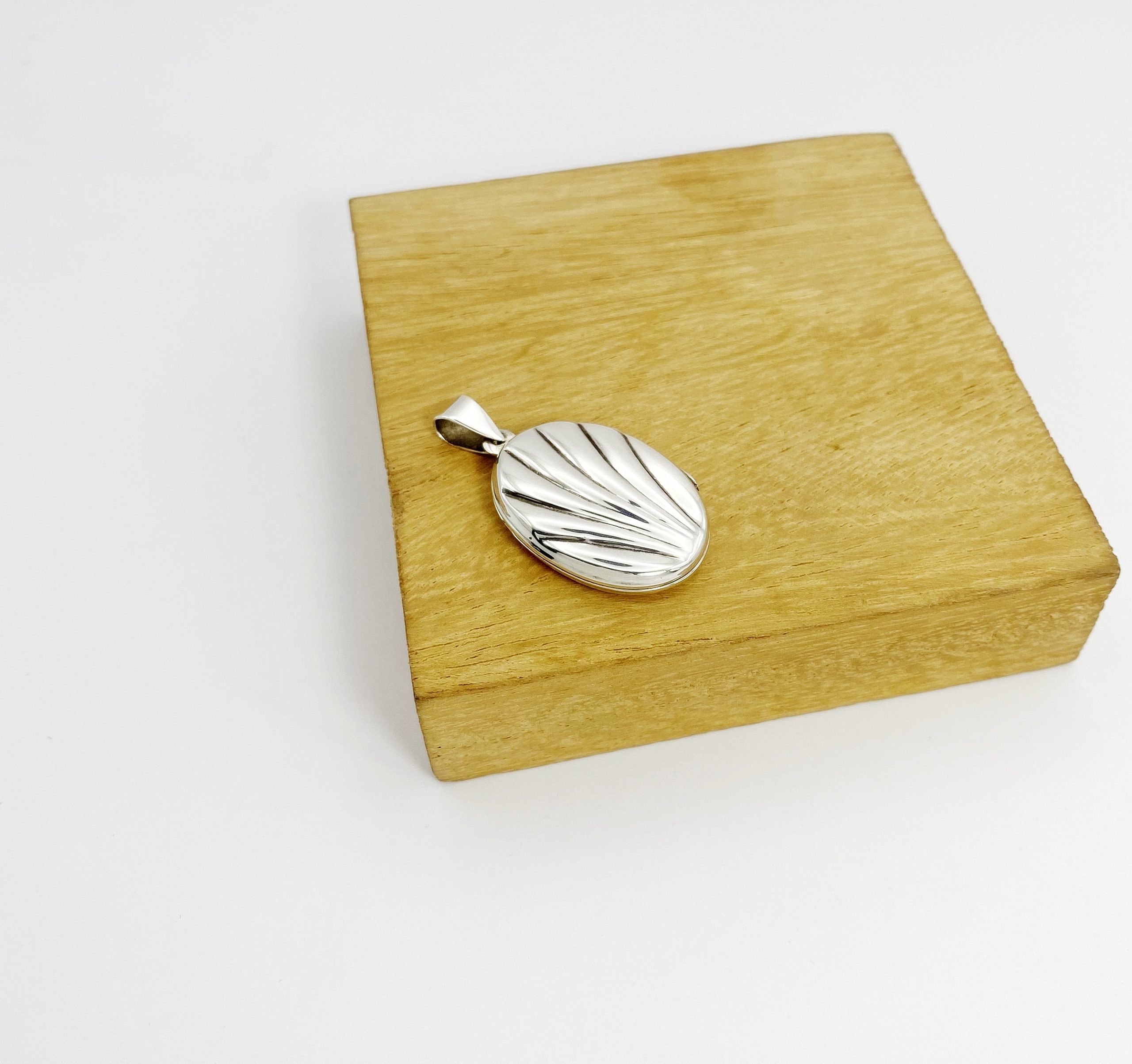 Side view of a closed shell-textured silver locket highlighting the secure clasp and loop on a wooden stand.